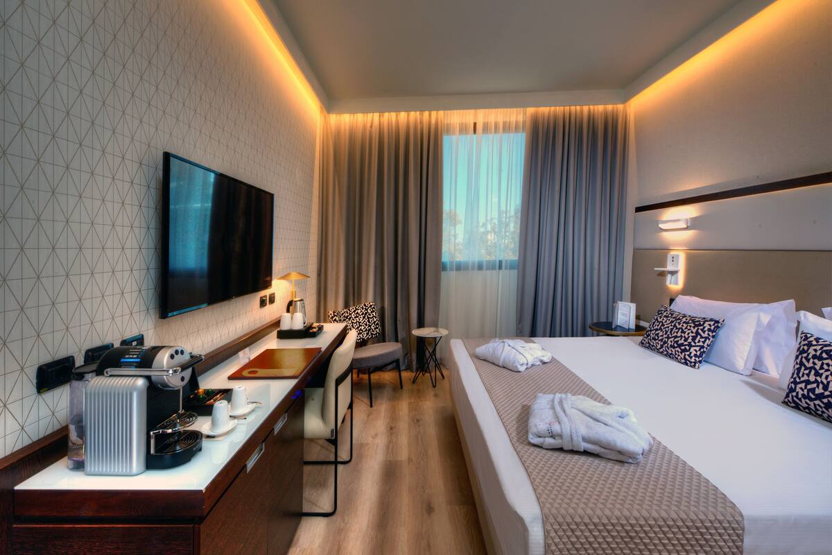 A modern hotel room with a large bed, a desk with a coffee machine, a flat-screen TV, and a view of the city through the window.