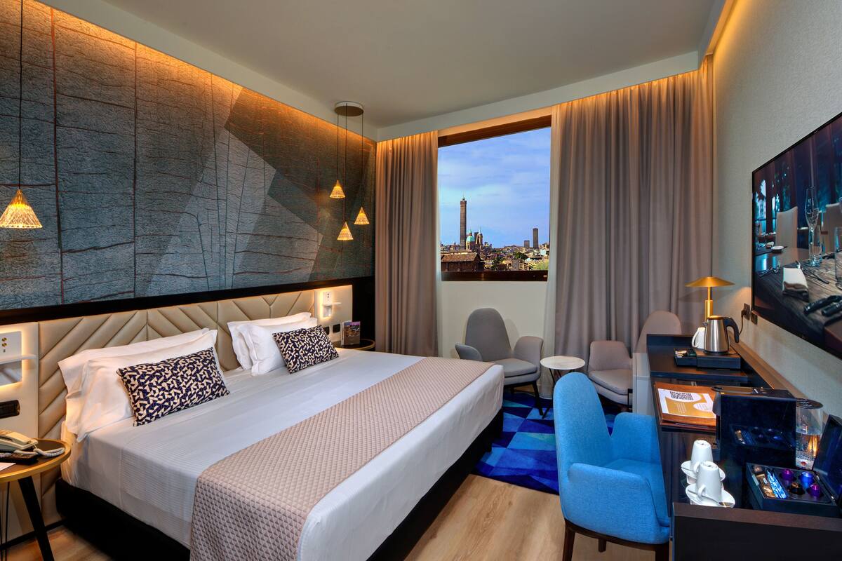 A modern hotel room with a large bed, a desk with a coffee machine, a chair, and a view of a cityscape.