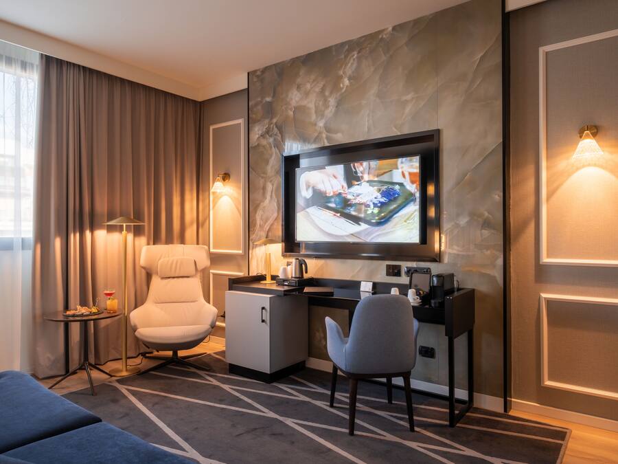 A hotel room with a TV, a desk, a chair, a sofa, a lamp, and a small table with a drink.