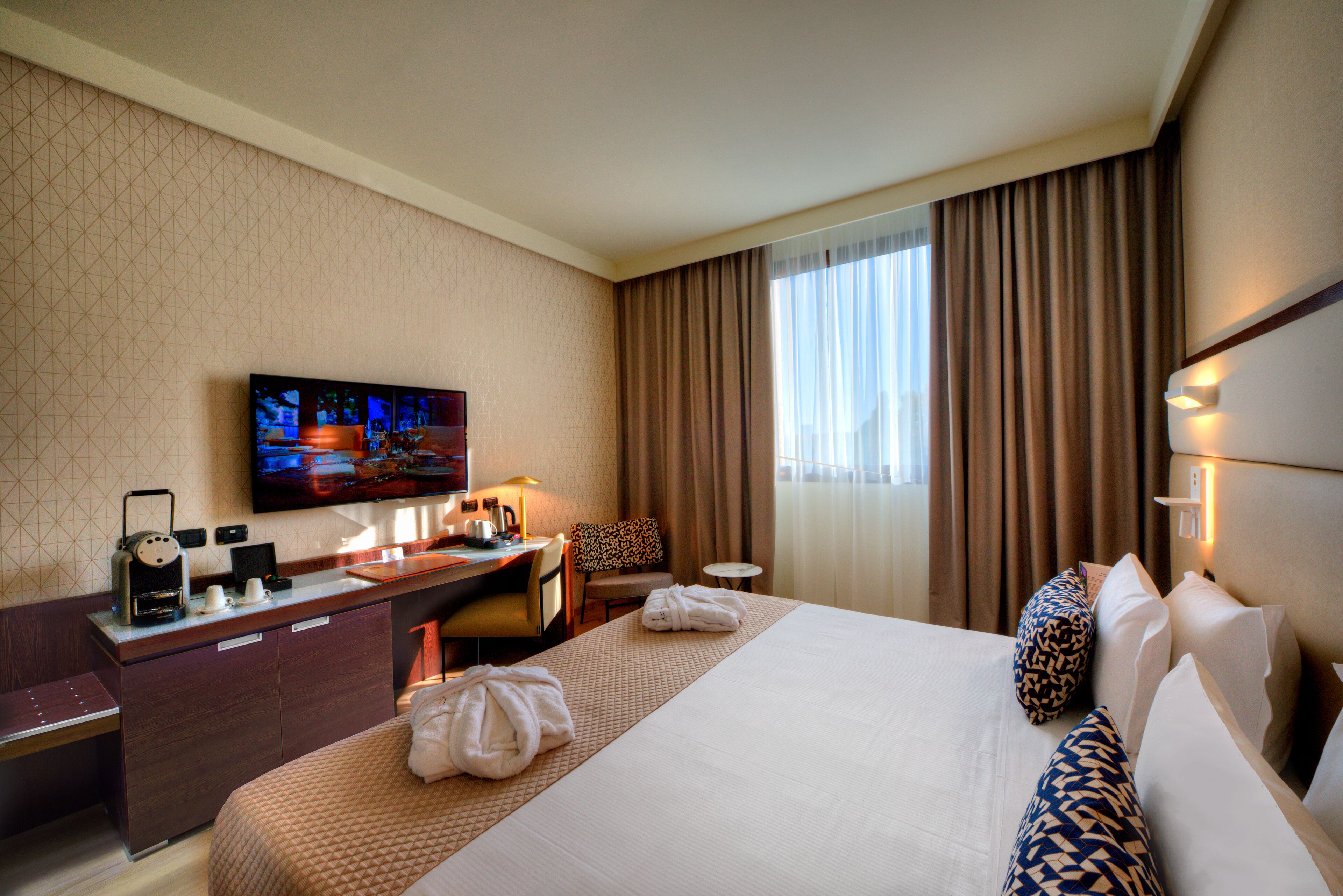 Quarto de hotel com uma cama grande, uma escrivaninha com televisão de tela plana, cafeteira e uma cadeira.