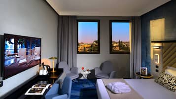 Habitación de hotel con una cama, dos sillones, una mesita con una lámpara, un televisor mostrando una escena de restaurante, y dos ventanas con vistas a la ciudad.