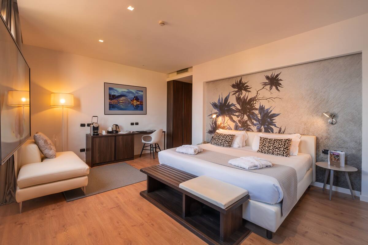 A modern hotel room with a large bed, a desk, a chair, and a sofa.