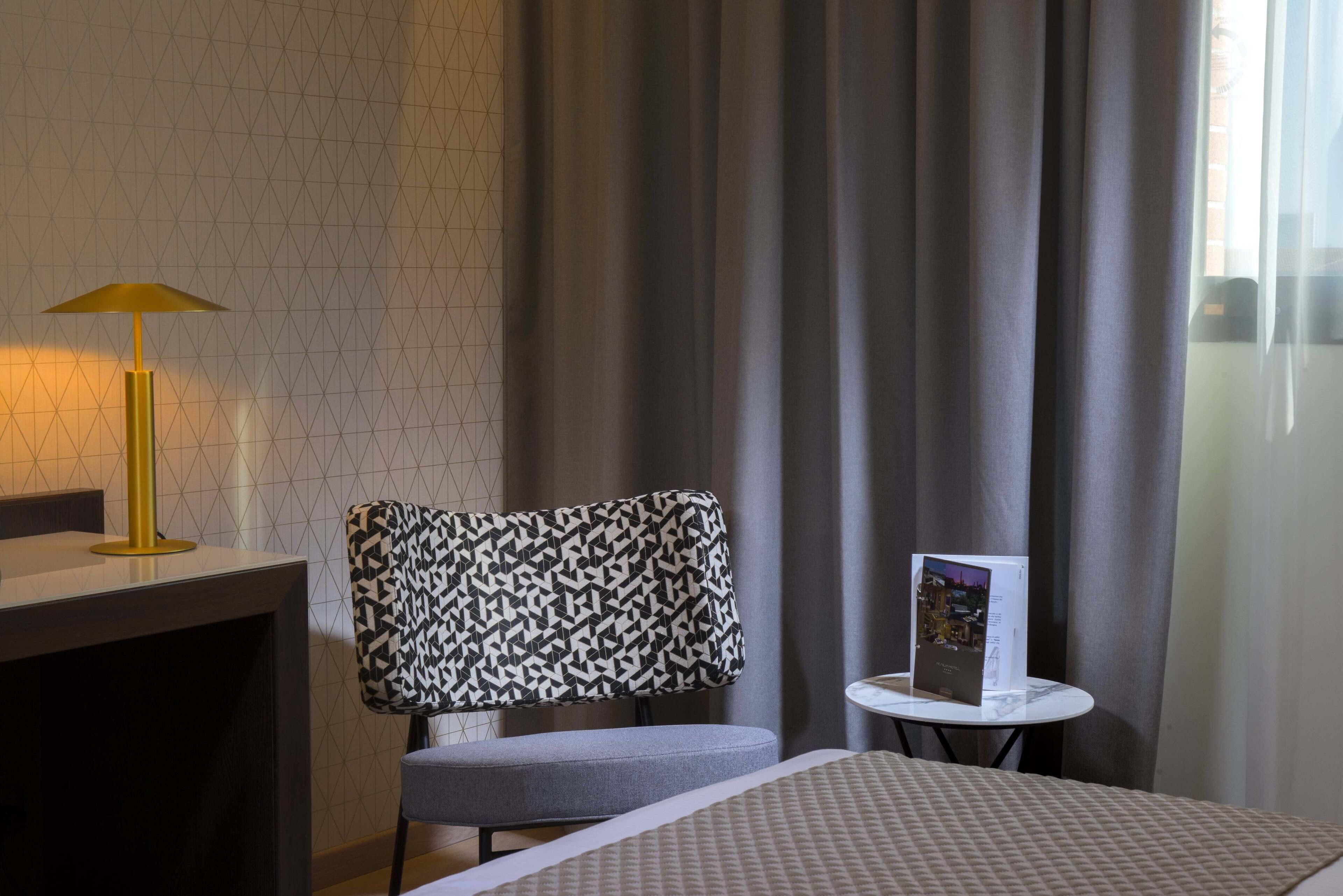 Ein Hotelzimmer mit einem gemusterten Sessel, einem kleinen Tisch mit einer Broschüre und einer Lampe.