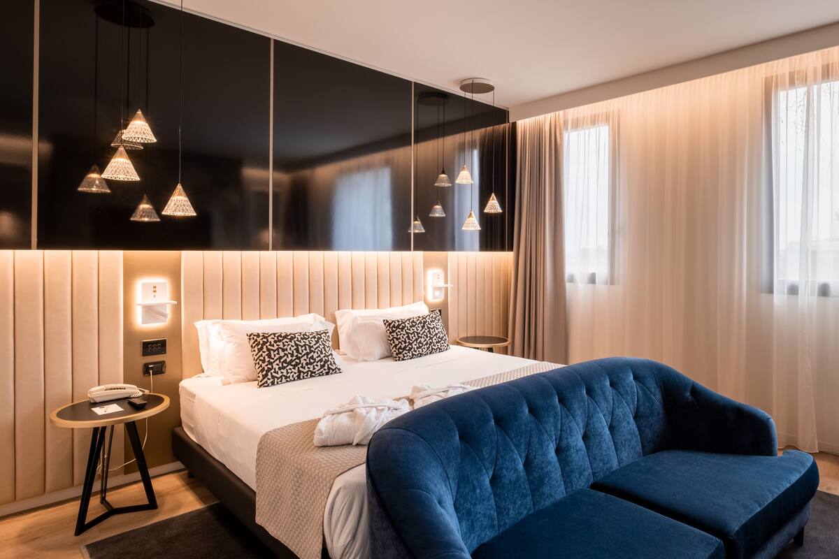 A modern hotel room with a large bed, a blue sofa, pendant lights, a bedside table, and a view of the outdoors through sheer curtains.