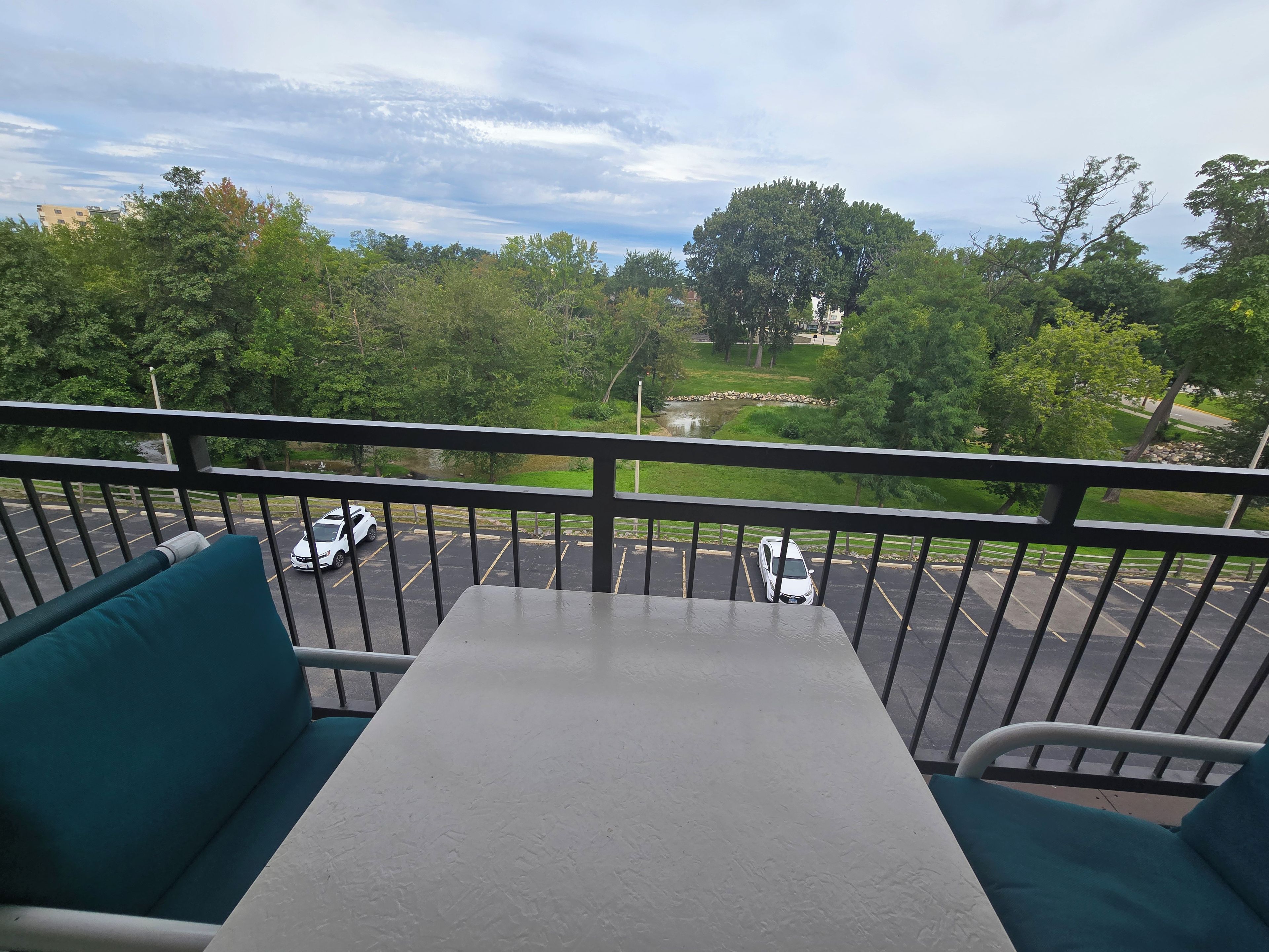 Un balcon avec une table et des chaises, donnant sur un parking et sur un espace verdoyant.