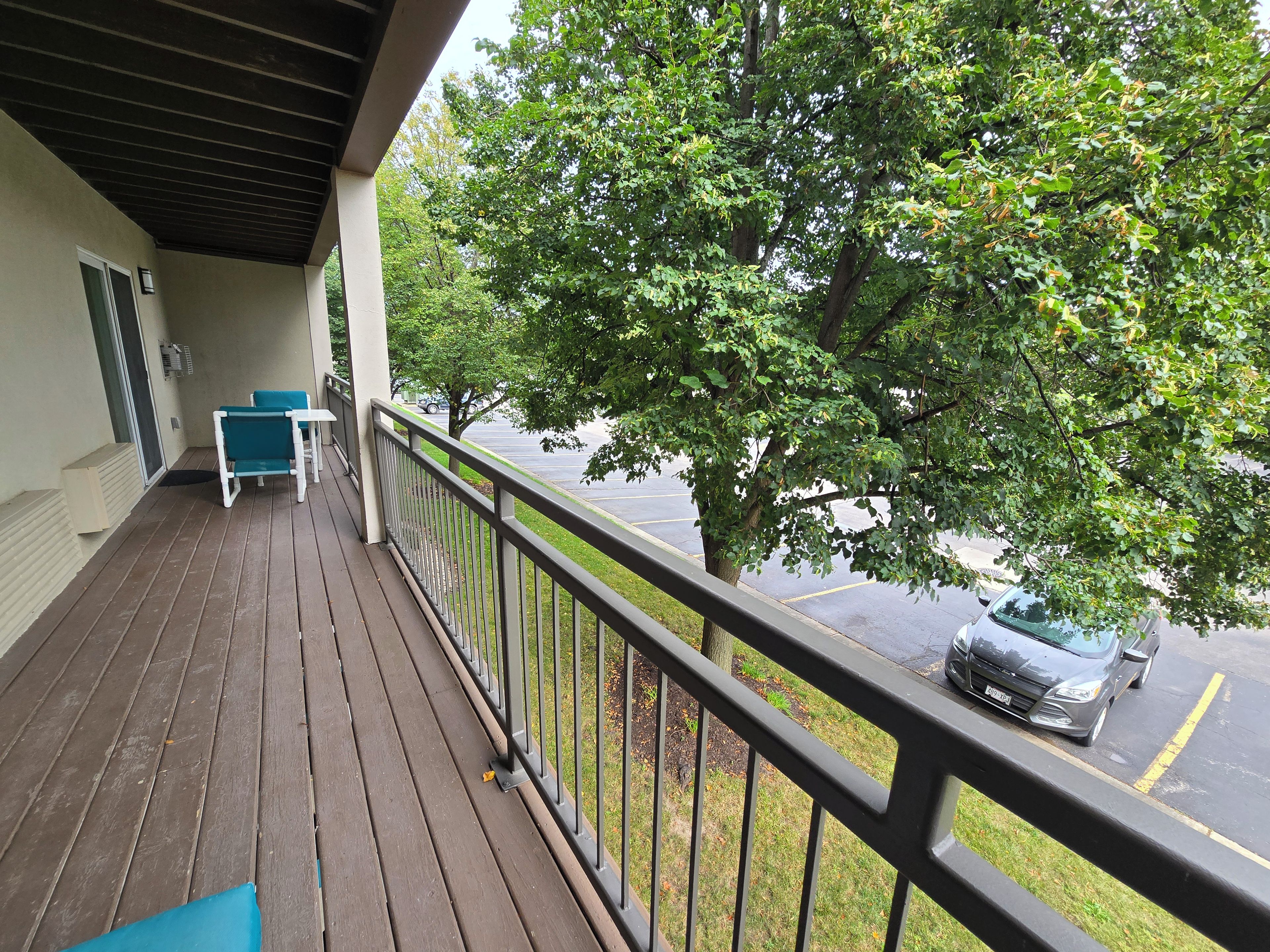 Um deck de madeira com guarda-corpo, uma mesa e cadeiras, com vista para um estacionamento e uma árvore.