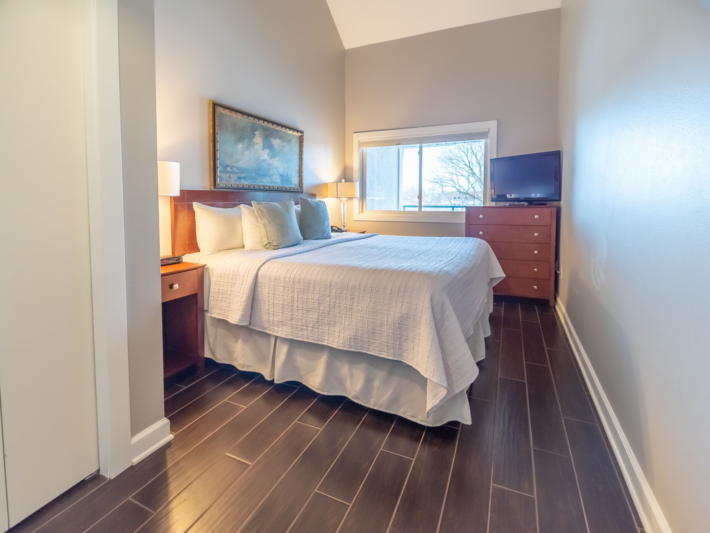 A bedroom with a bed, nightstands, a dresser, a television, and a window with a view of trees.