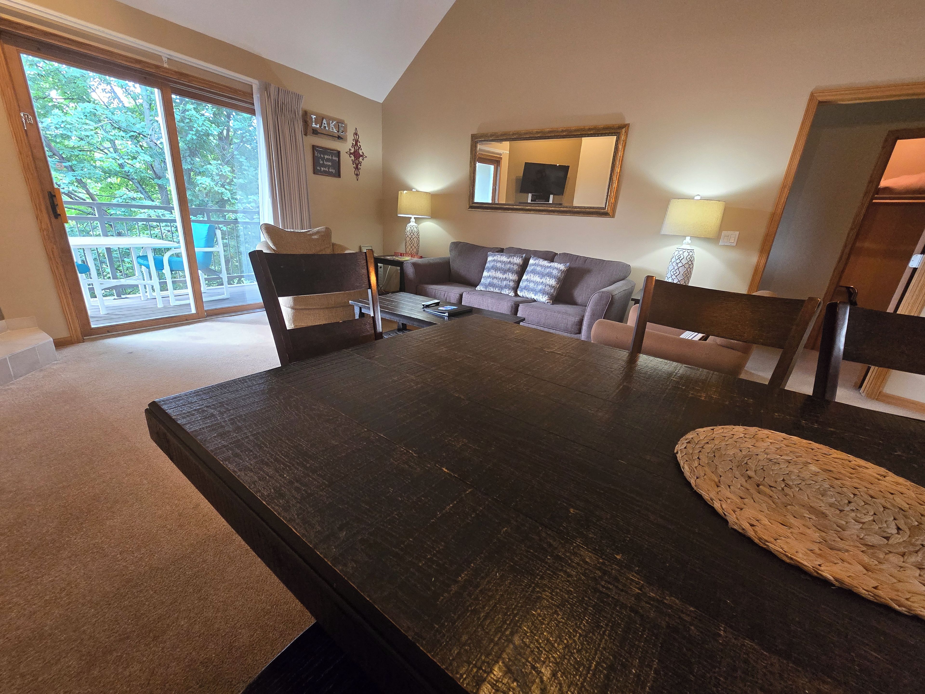 Een eethoek met een donker houten tafel, stoelen en een bank met kussens. Er is een groot raam met uitzicht op groen.