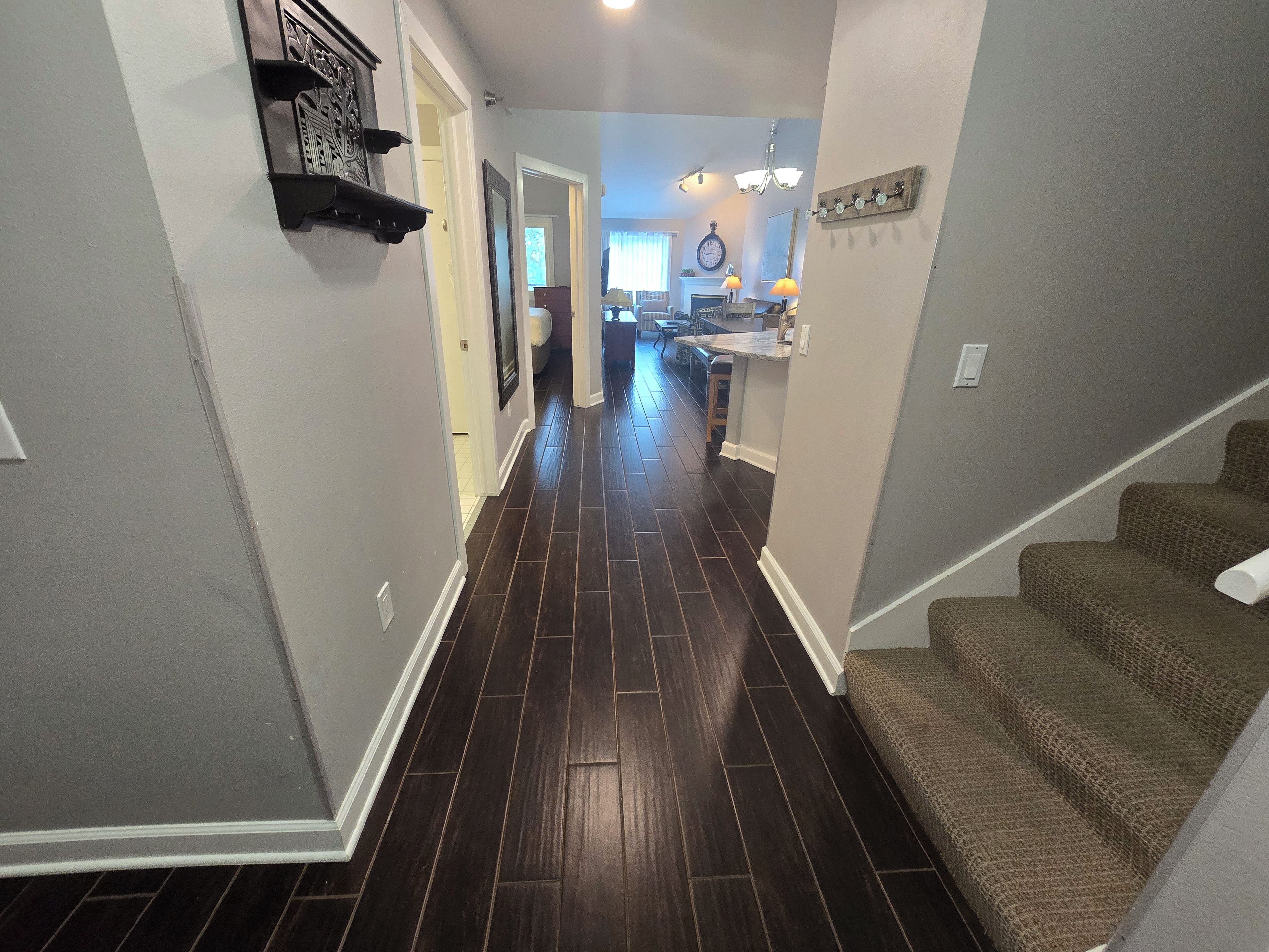 A hallway with dark wooden flooring, a staircase on the right, and a kitchen area in the background.