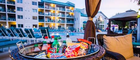 A wicker basket filled with snacks and drinks, lounge chairs, and a poolside view of a hotel with multiple balconies and a covered area.