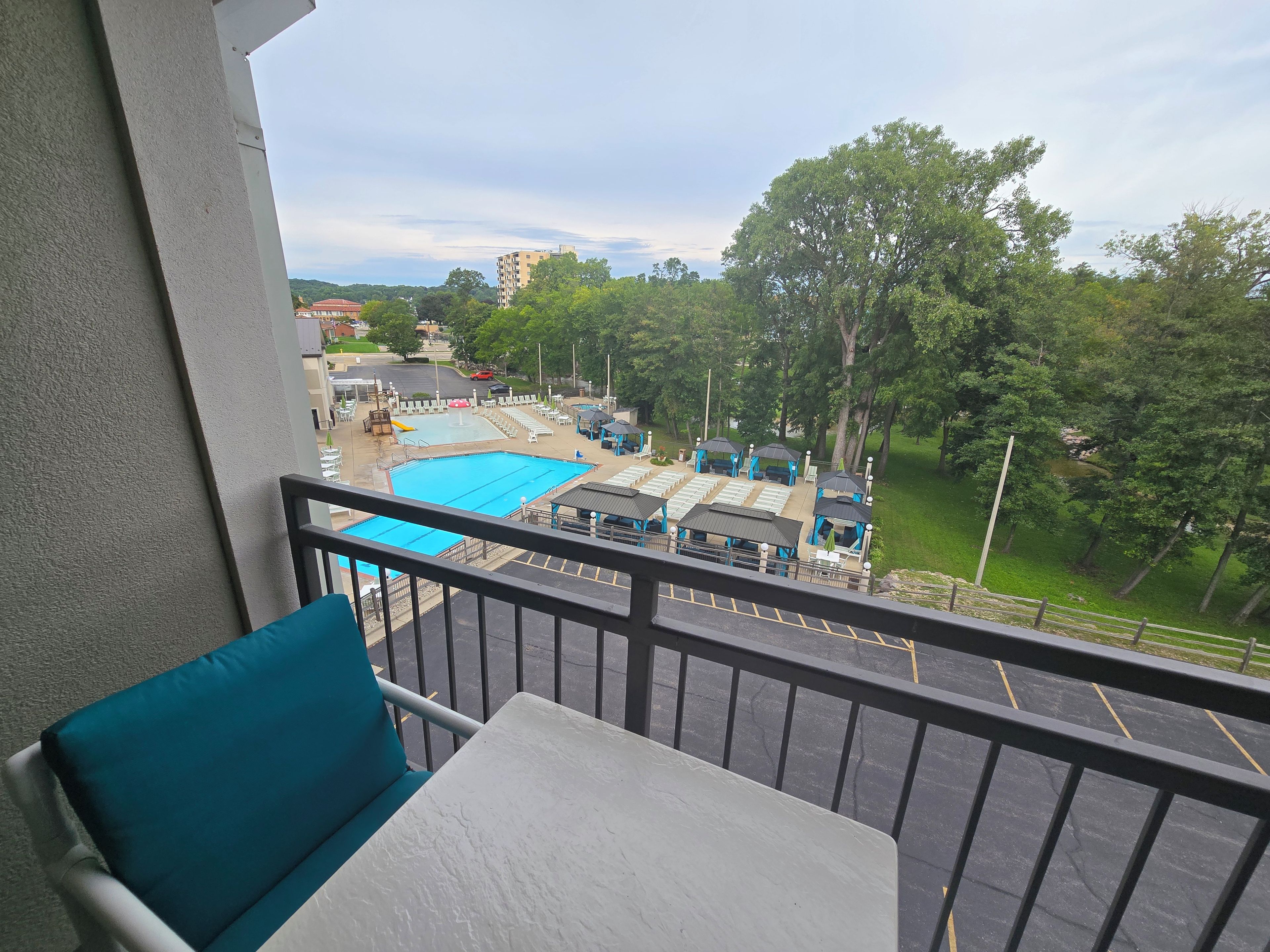 A balcony with a view of a swimming pool, lounge chairs, and a parking lot.