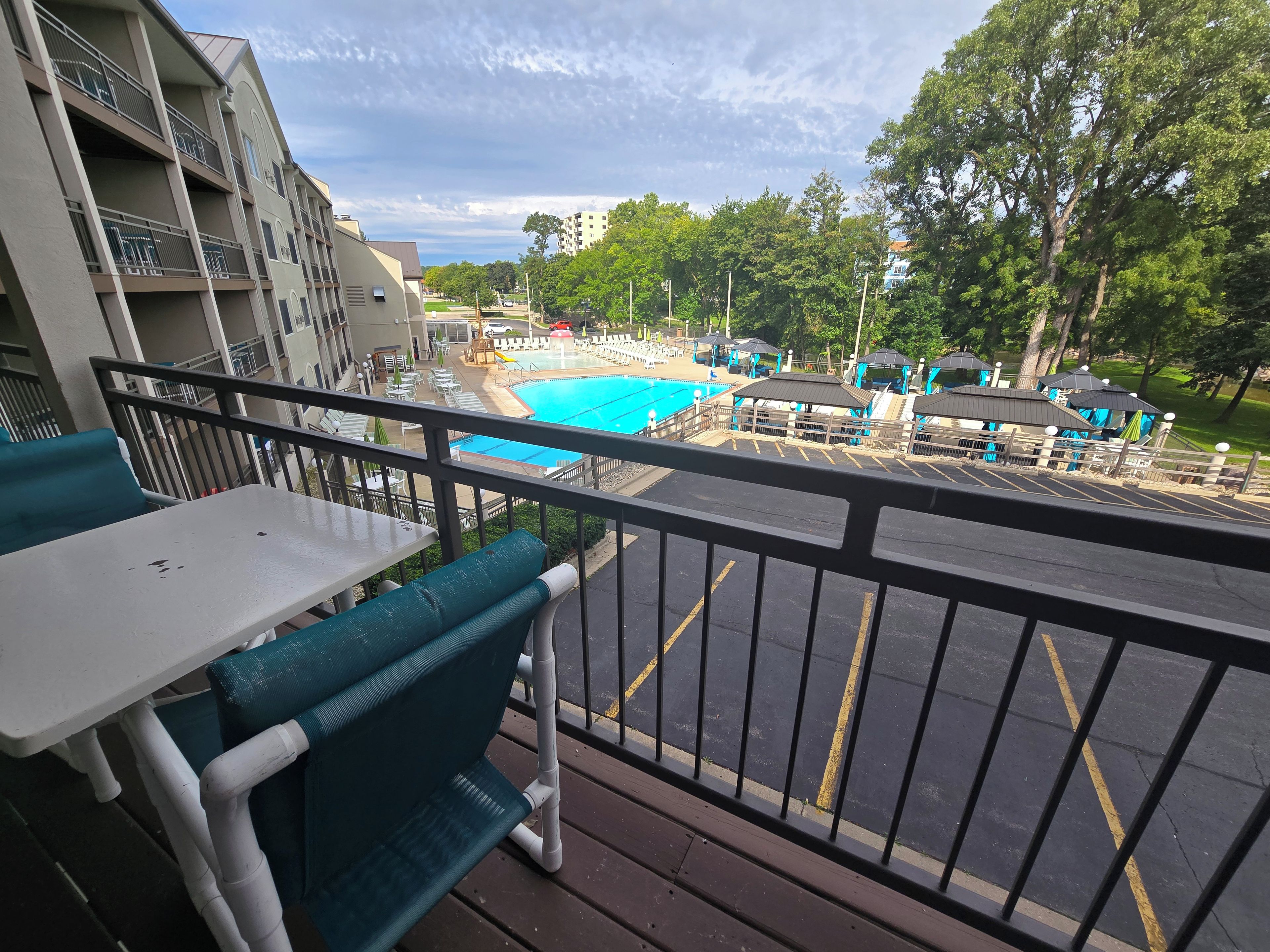 A balcony with a view of a swimming pool, outdoor seating, and a parking area.