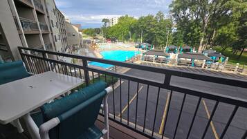 A balcony with a view of a swimming pool, outdoor seating, and a parking area.