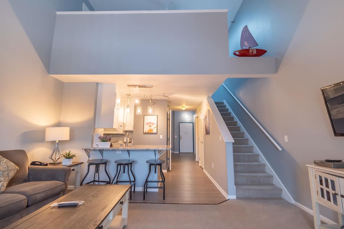 Un salon moderne avec un canapé, une table basse et des tabourets de bar. Un escalier mène à un espace cuisine doté d’un éclairage suspendu.