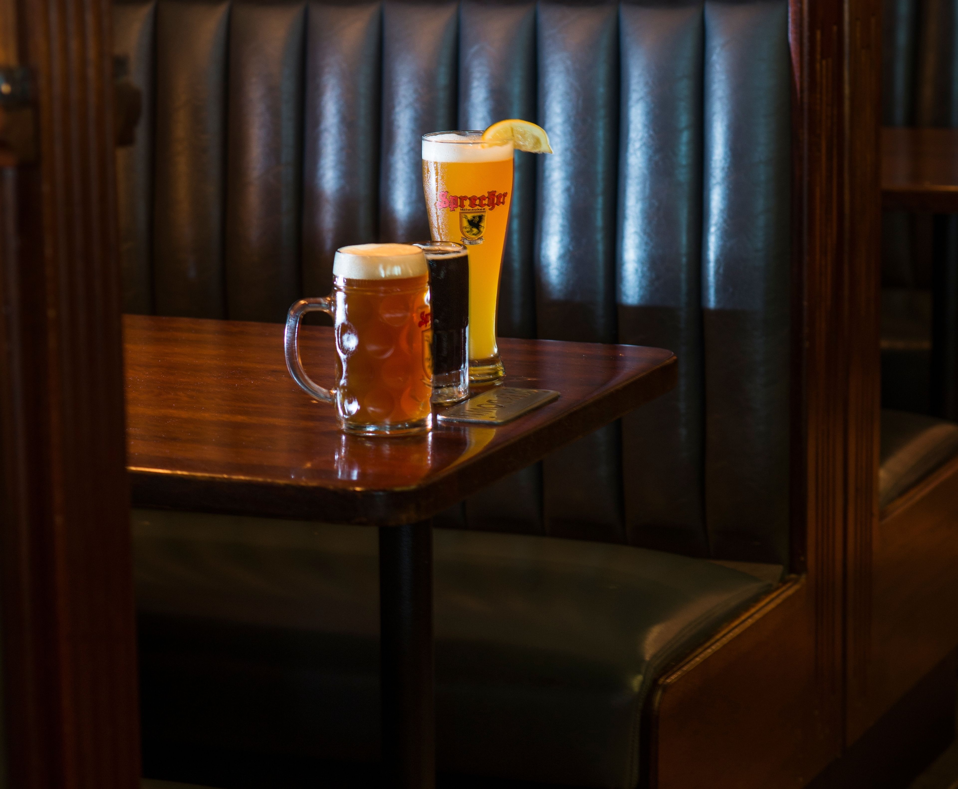 Une table en bois, sur laquelle sont posés deux chopes de bière et un grand verre de bière garni d’une tranche de citron, le tout placé devant une banquette en cuir foncé.