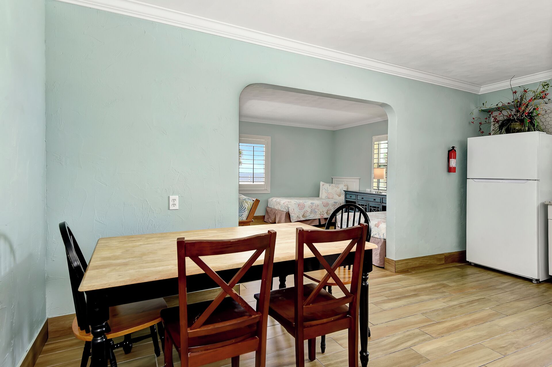 Un coin repas avec une table et des chaises en bois, un réfrigérateur blanc et une cuisine visible à travers une arche.