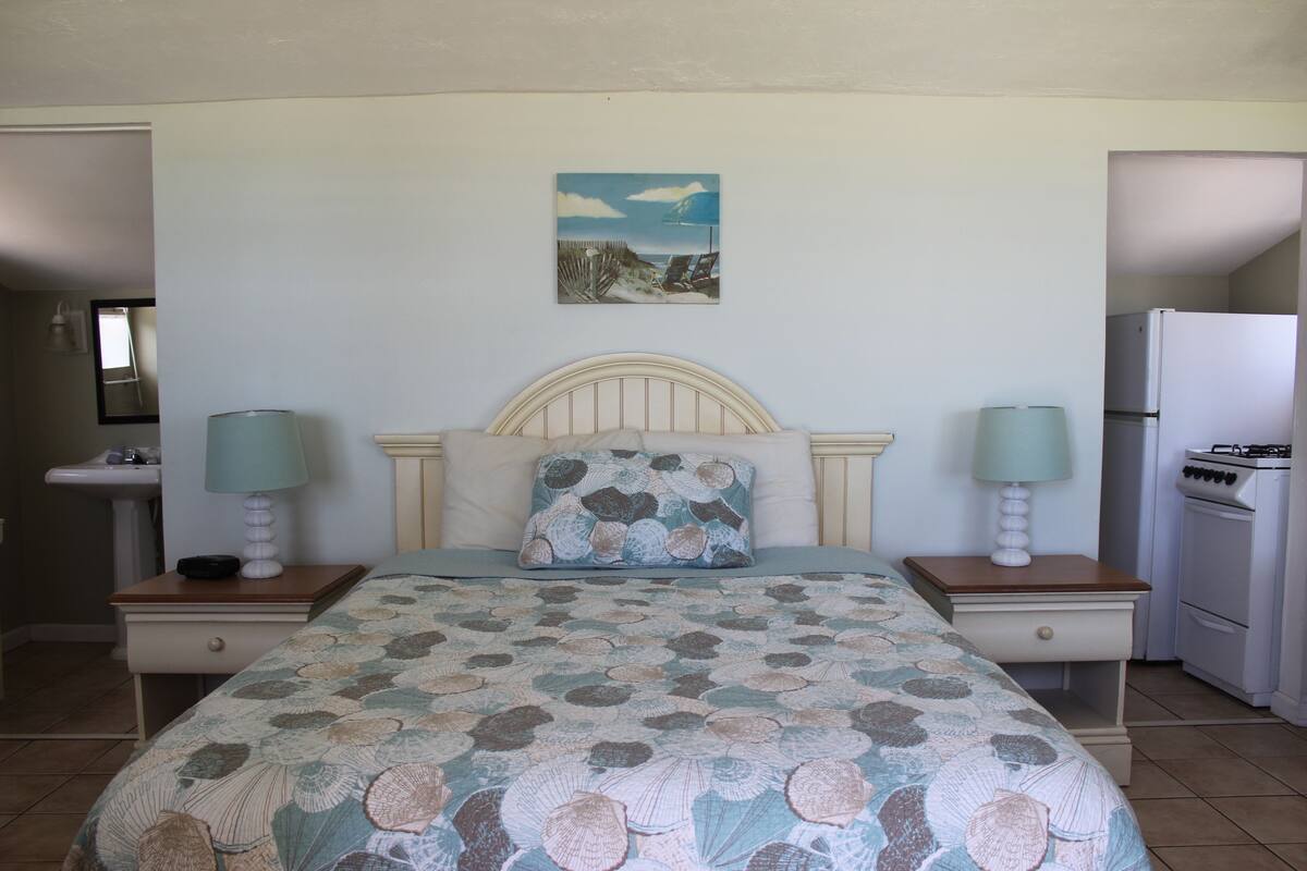 Un dormitorio con cama, dos mesitas de noche con lámparas, una mesita auxiliar, un cuadro en la pared y una zona de cocina con refrigerador y estufa.