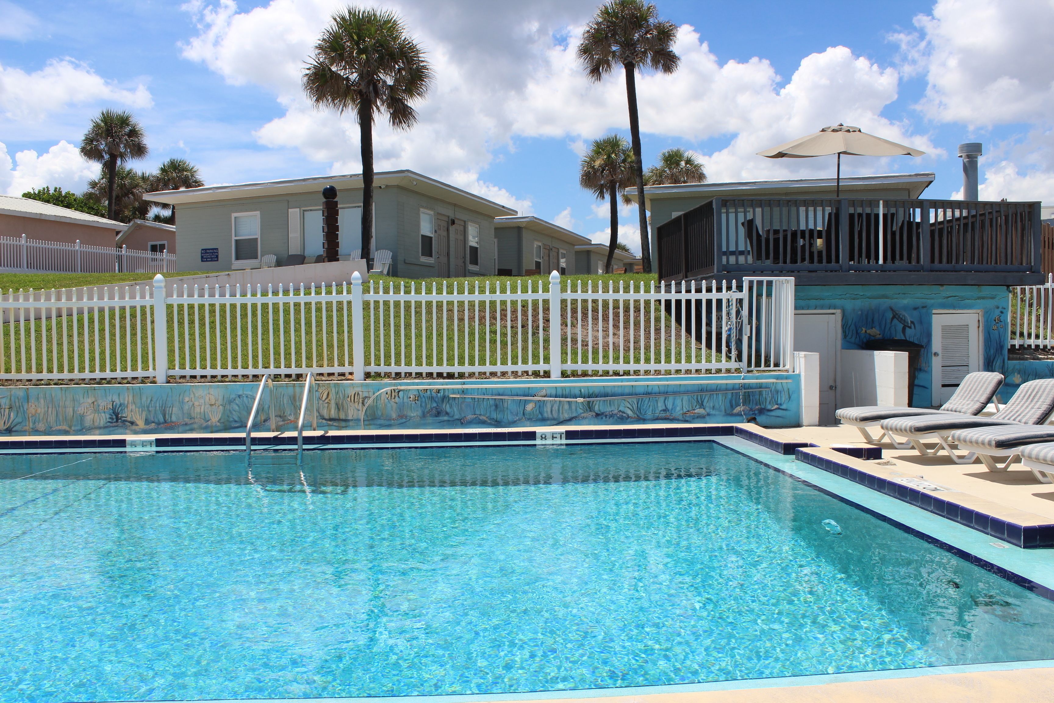 A swimming pool with a white fence, lounge chairs, and palm trees in the background.