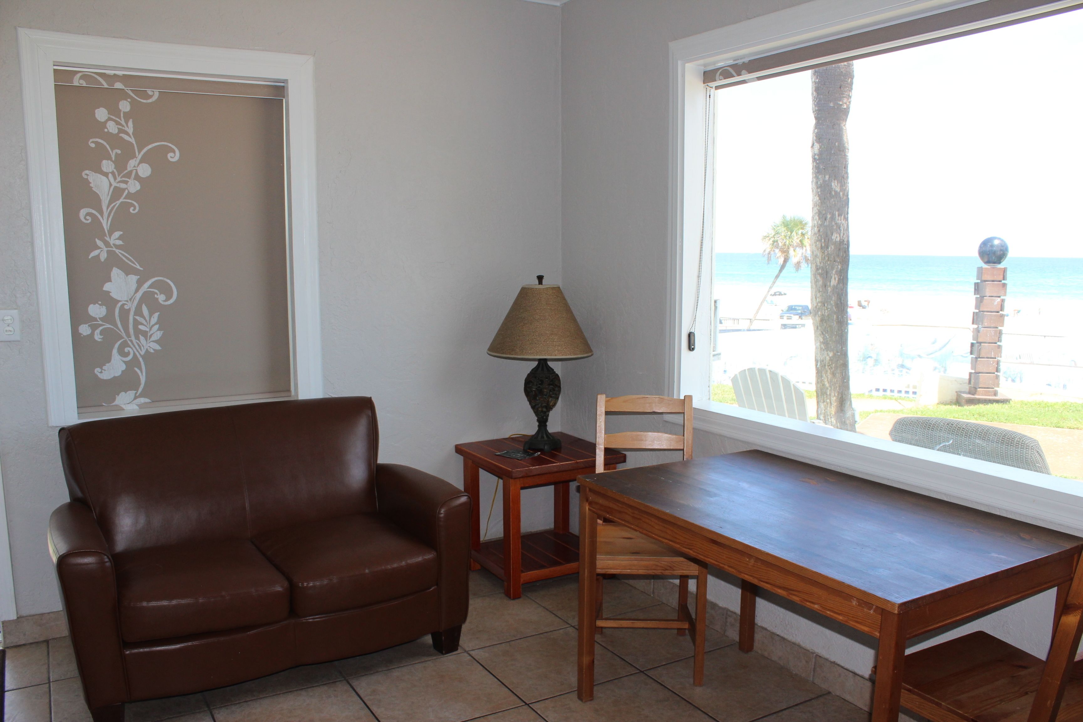 A room with a brown leather sofa, a wooden table, a lamp, and a view of the beach through a large window.