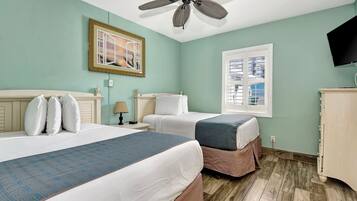 A hotel room with two beds, a ceiling fan, a window with blinds, a television, and a wooden floor.