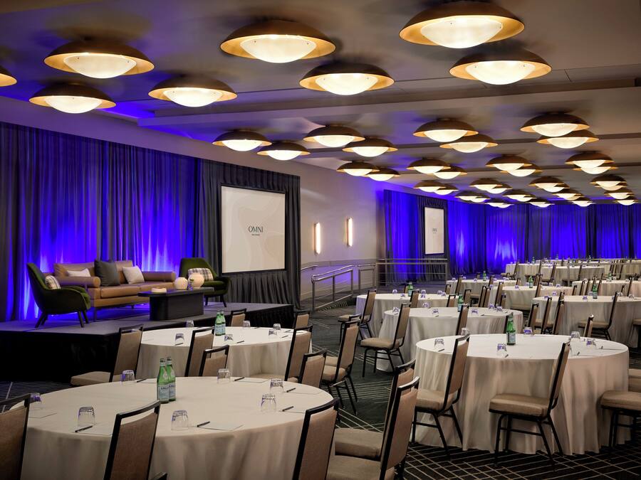 Un salón de conferencias con varias mesas redondas, sillas y un escenario con una pantalla.