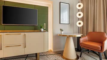 A hotel room with a flat-screen TV mounted on a green panel, a wooden cabinet with drawers, a round table with a potted plant, a leather chair, and a floor lamp with three lights.