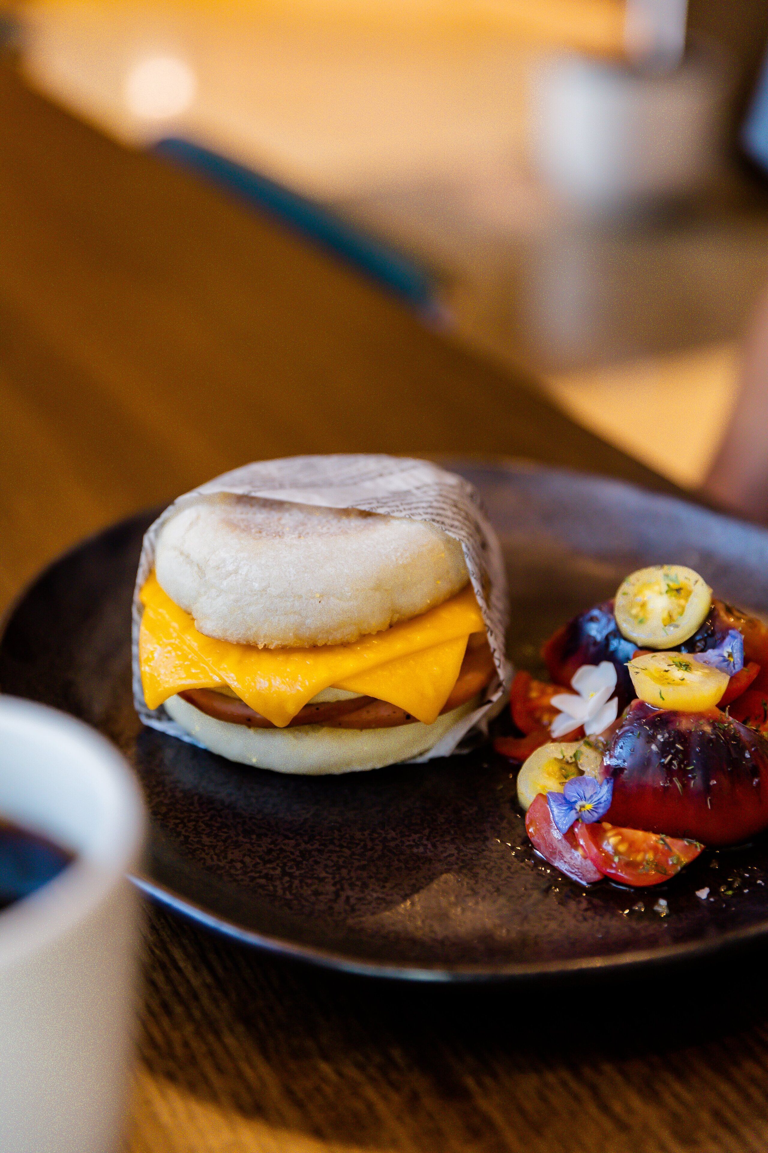 A breakfast sandwich on a plate with a side of sliced tomatoes and a small flower.
