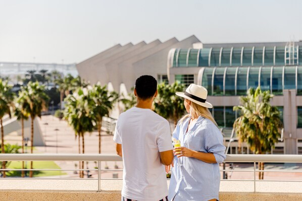 A couple standing on a balcony overlooking a modern building with palm trees in the background.