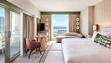 A hotel room with a large bed, a sofa, a TV, a small table, and a view of a cityscape through the window.