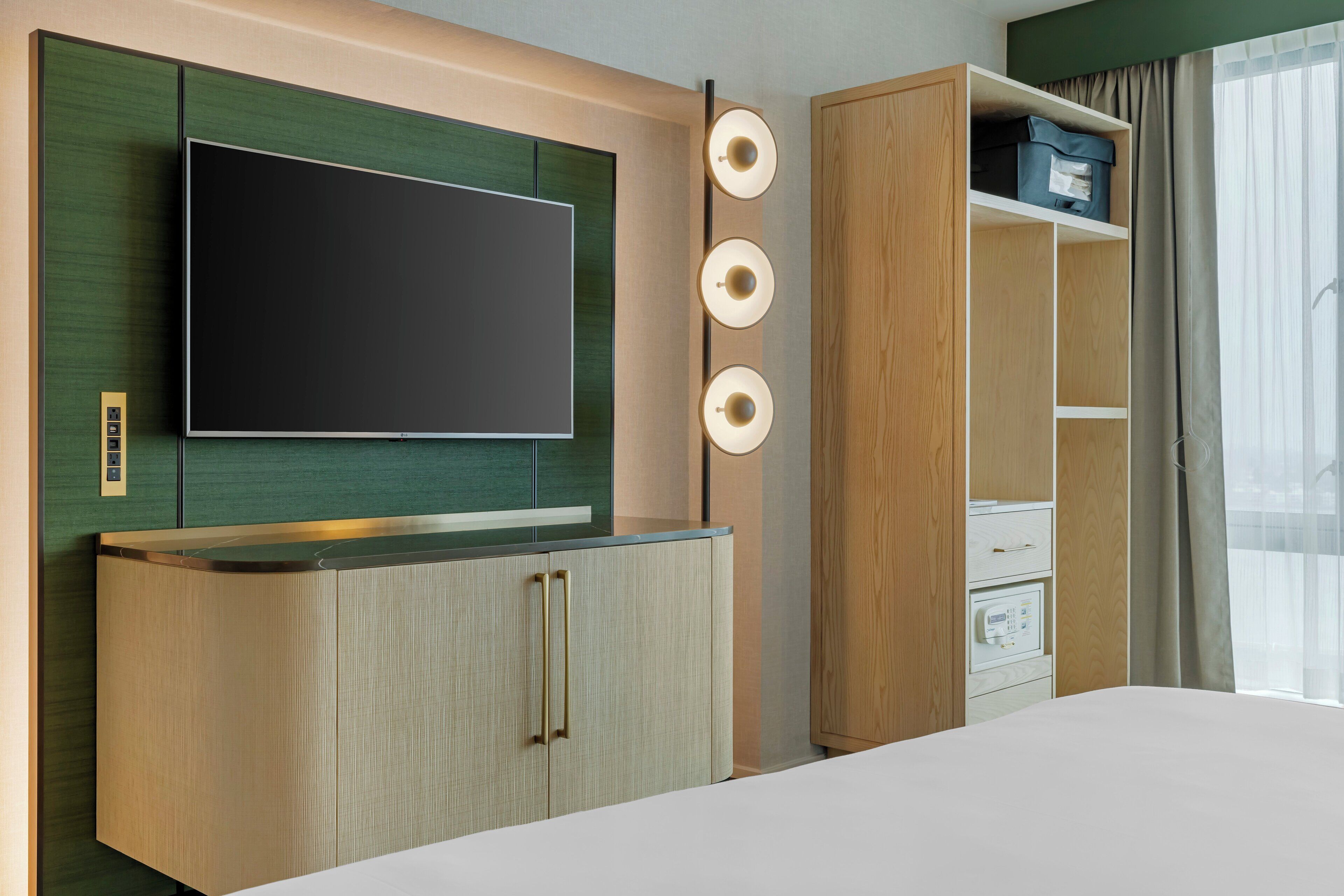 A modern hotel room with a large flat-screen TV mounted on a green panel, a wooden cabinet with drawers, a bedside table, and a window with curtains.