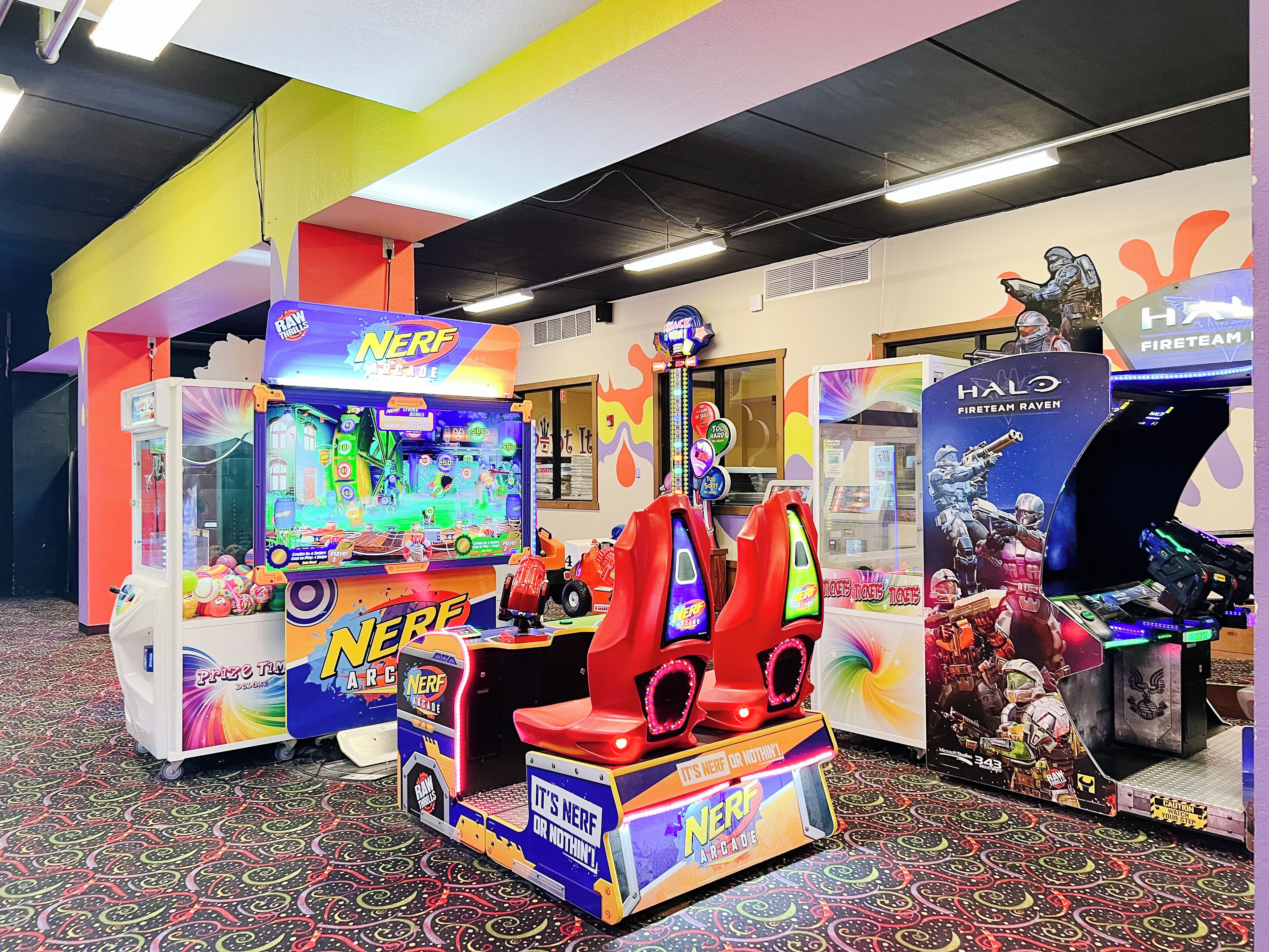 Et farverigt arkadeområde med forskellige spillemaskiner, herunder en 'Nerf Arcade' og et 'Halo Fireteam Raven'-spil.