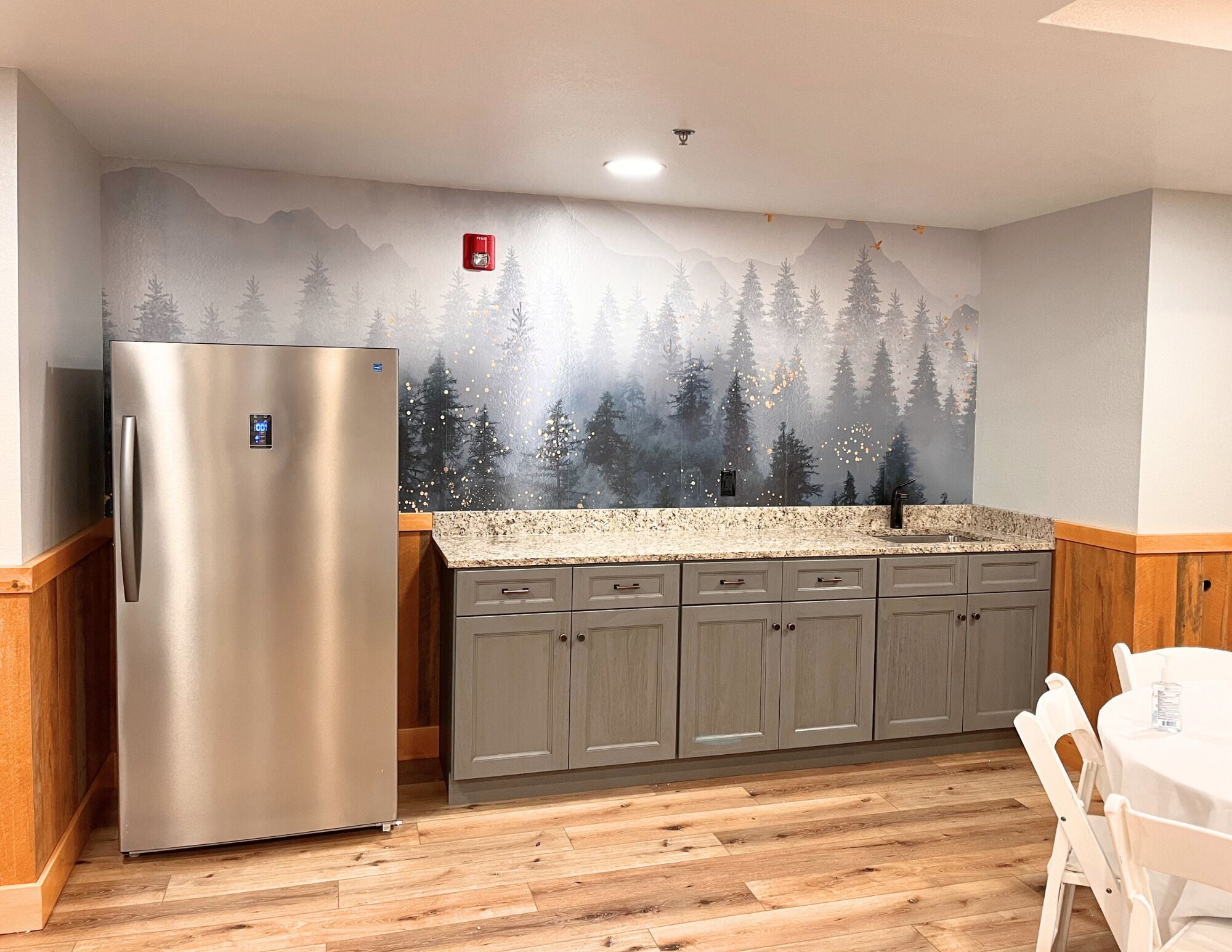 A kitchen with a stainless steel refrigerator, grey cabinets, a granite countertop, and a forest-themed wall mural.