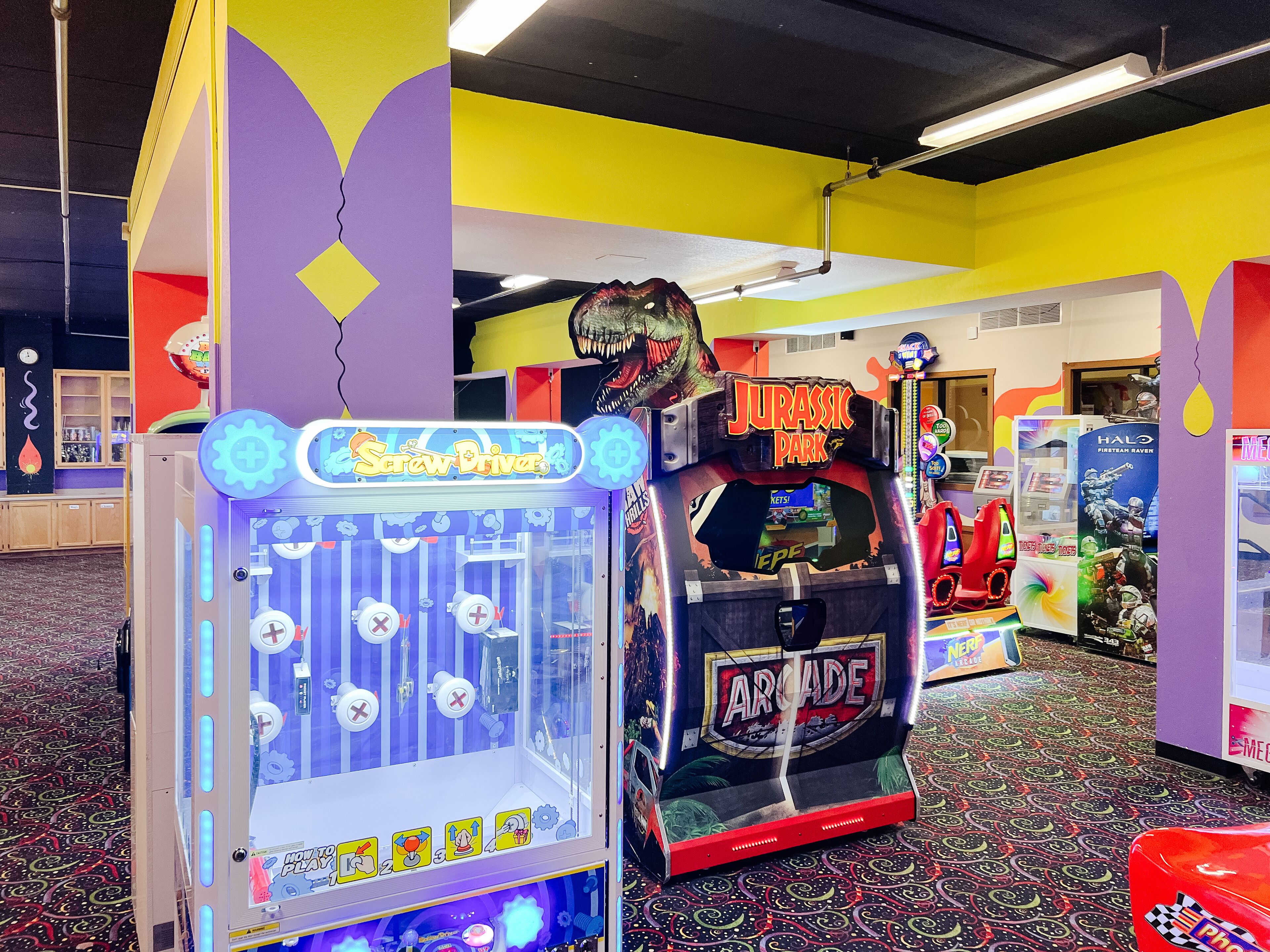A brightly lit arcade with various games, including a 'Jurassic Park' themed machine and a claw game.