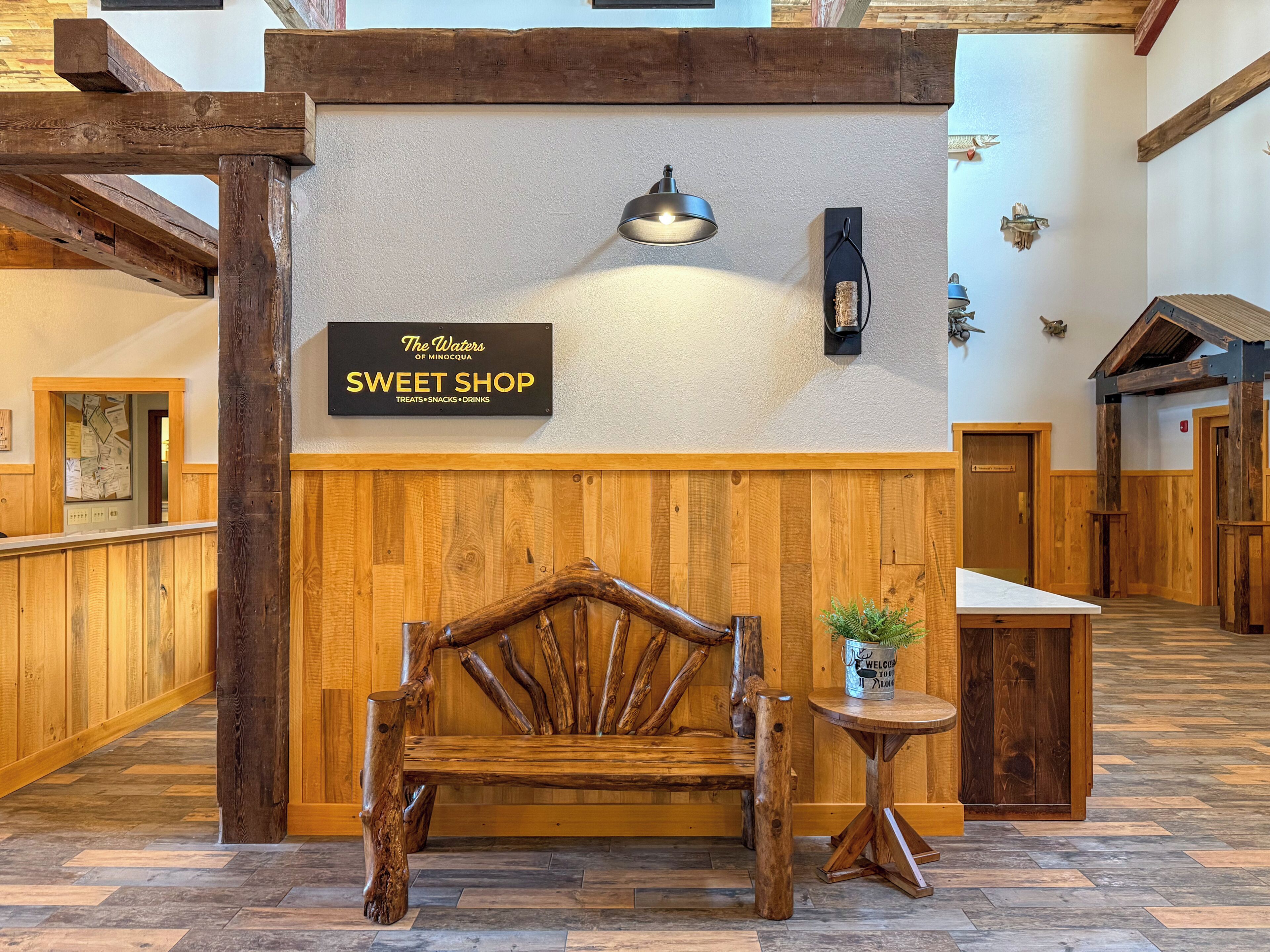 Un banc en bois avec une pancarte au-dessus, une petite table avec une plante et un mur en bois orné d’une autre pancarte.