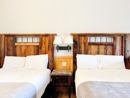 Una habitación con dos camas, cabeceros de madera, una mesita de noche con lámpara y un ventanal pequeño.
