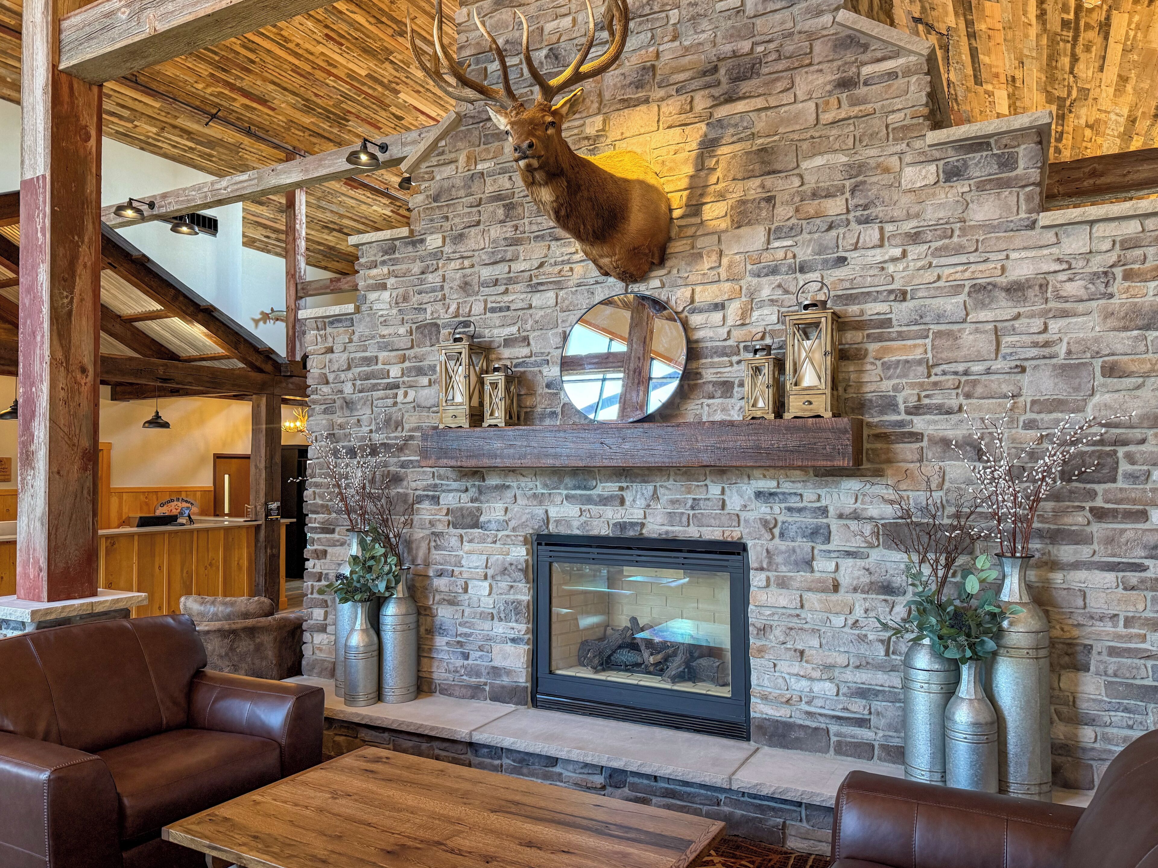 A rustic interior with a stone wall, a mounted deer head, a fireplace, leather seating, and metal vases.