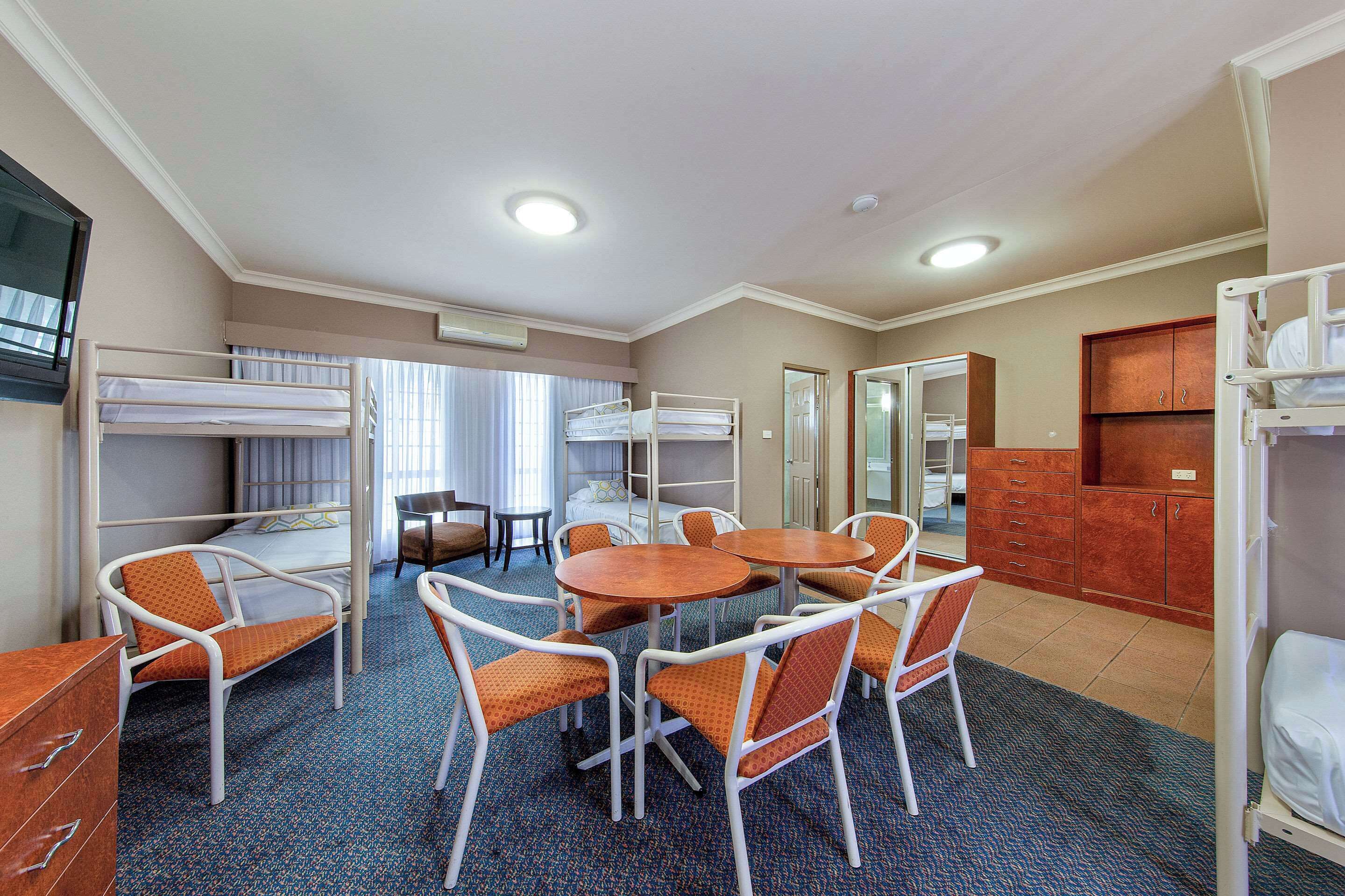 A hotel room with bunk beds, a dining table with chairs, a small kitchen area, and a window with curtains.