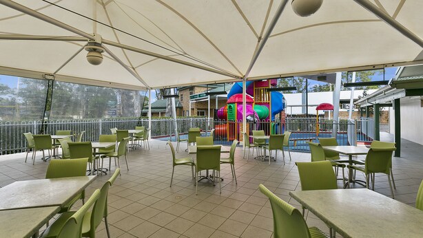 An outdoor patio area with green tables and chairs, a play structure in the background, and a view of a building across the railing.