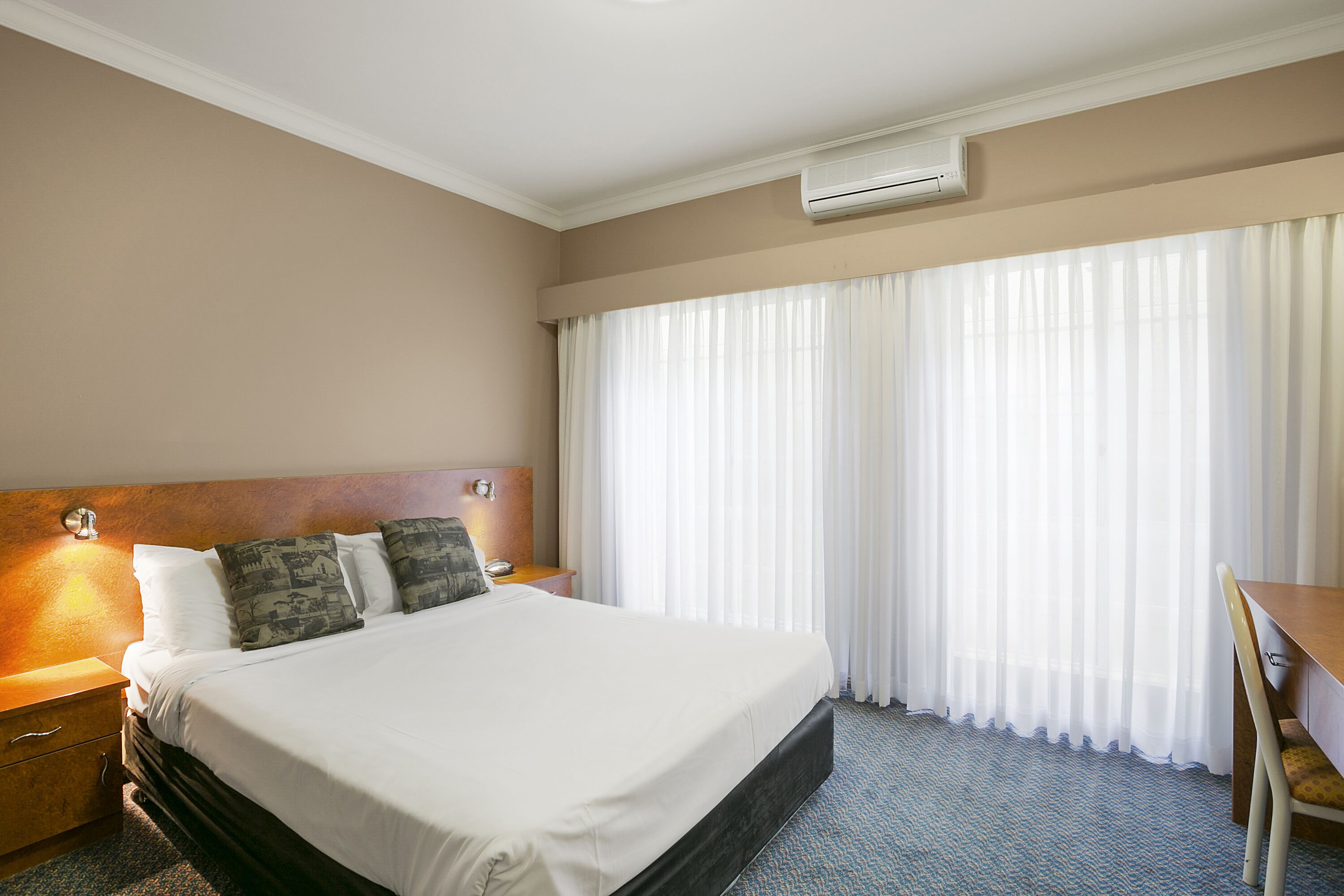 A hotel room with a large bed, a wooden headboard, a desk, and a window with sheer curtains.