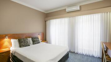 A hotel room with a large bed, a wooden headboard, a desk, and a window with sheer curtains.