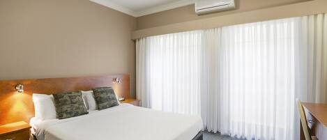 A hotel room with a large bed, a wooden headboard, a desk, and a window with sheer curtains.