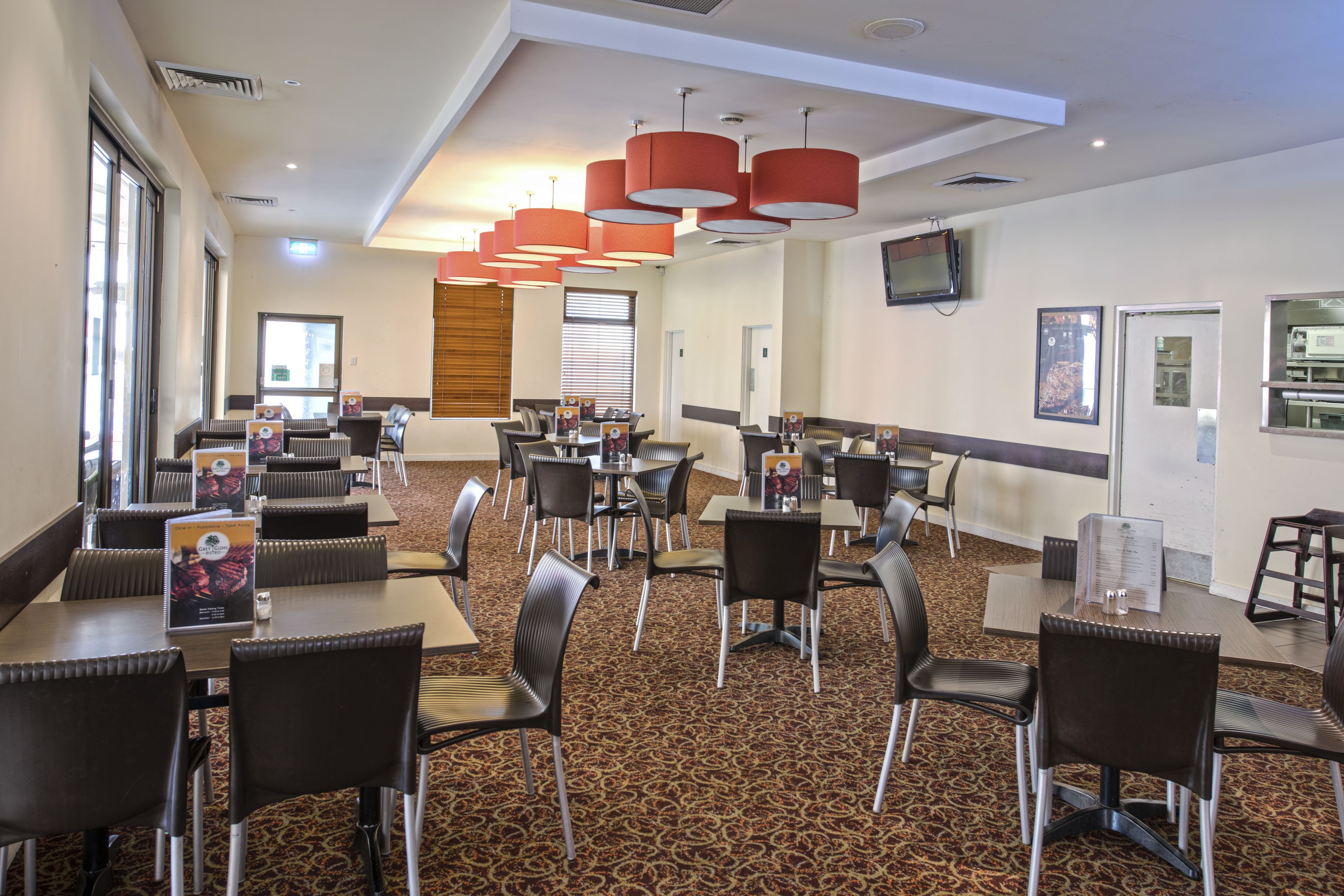A hotel dining area with multiple tables and chairs, a patterned carpet, and red pendant lights.