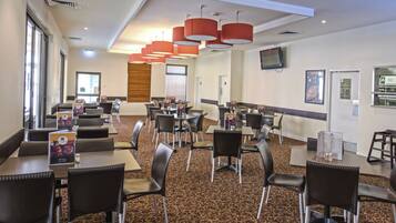 A hotel dining area with multiple tables and chairs, a patterned carpet, and red pendant lights.