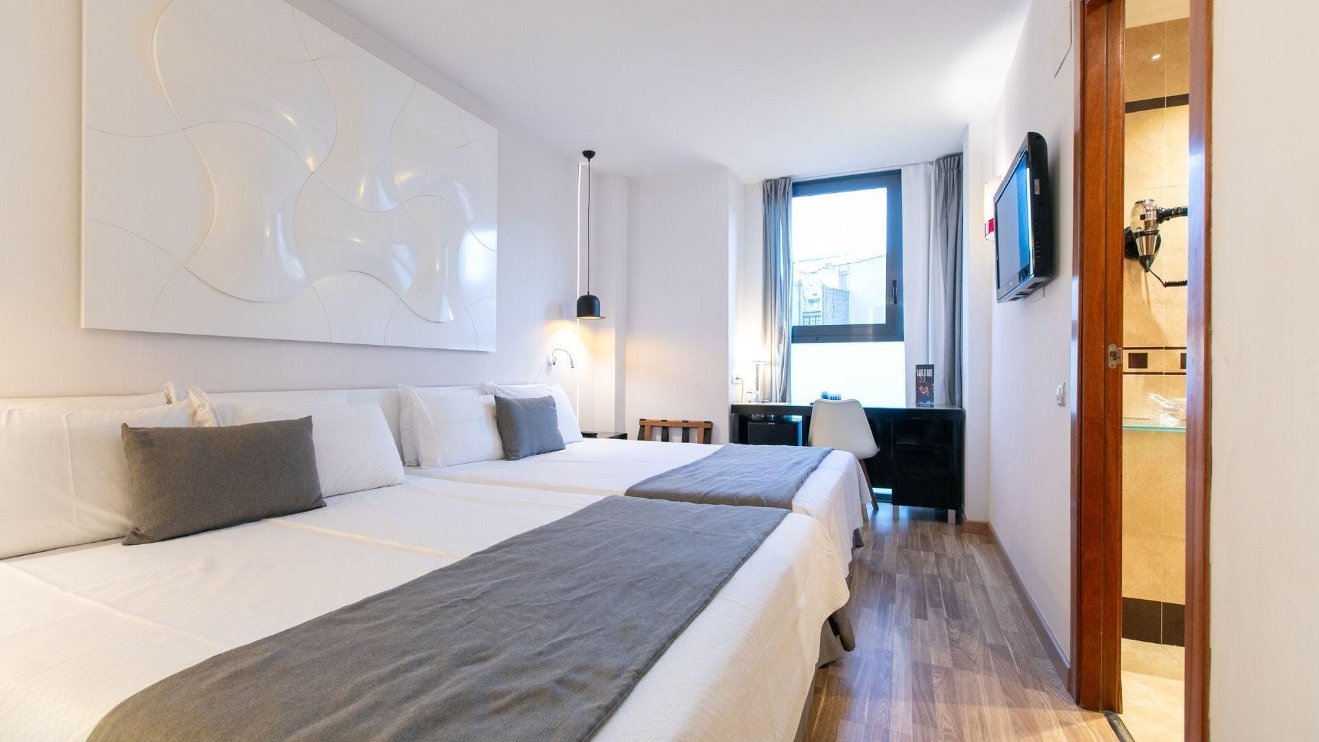Una habitació d'hotel amb dos llits, un quadre abstracte gran, una taula de treball amb una cadira, una televisió i una finestra amb cortines.