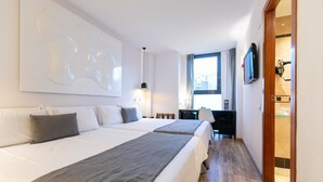 A hotel room with two beds, a desk, a TV, and a bathroom visible through an open door. - Evenia Rocafort (Barcelona)