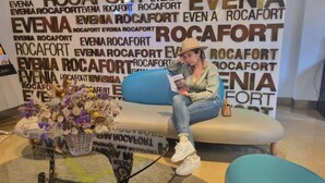A woman sitting on a blue sofa in a room with a backdrop of 'ROCAFORT' text.