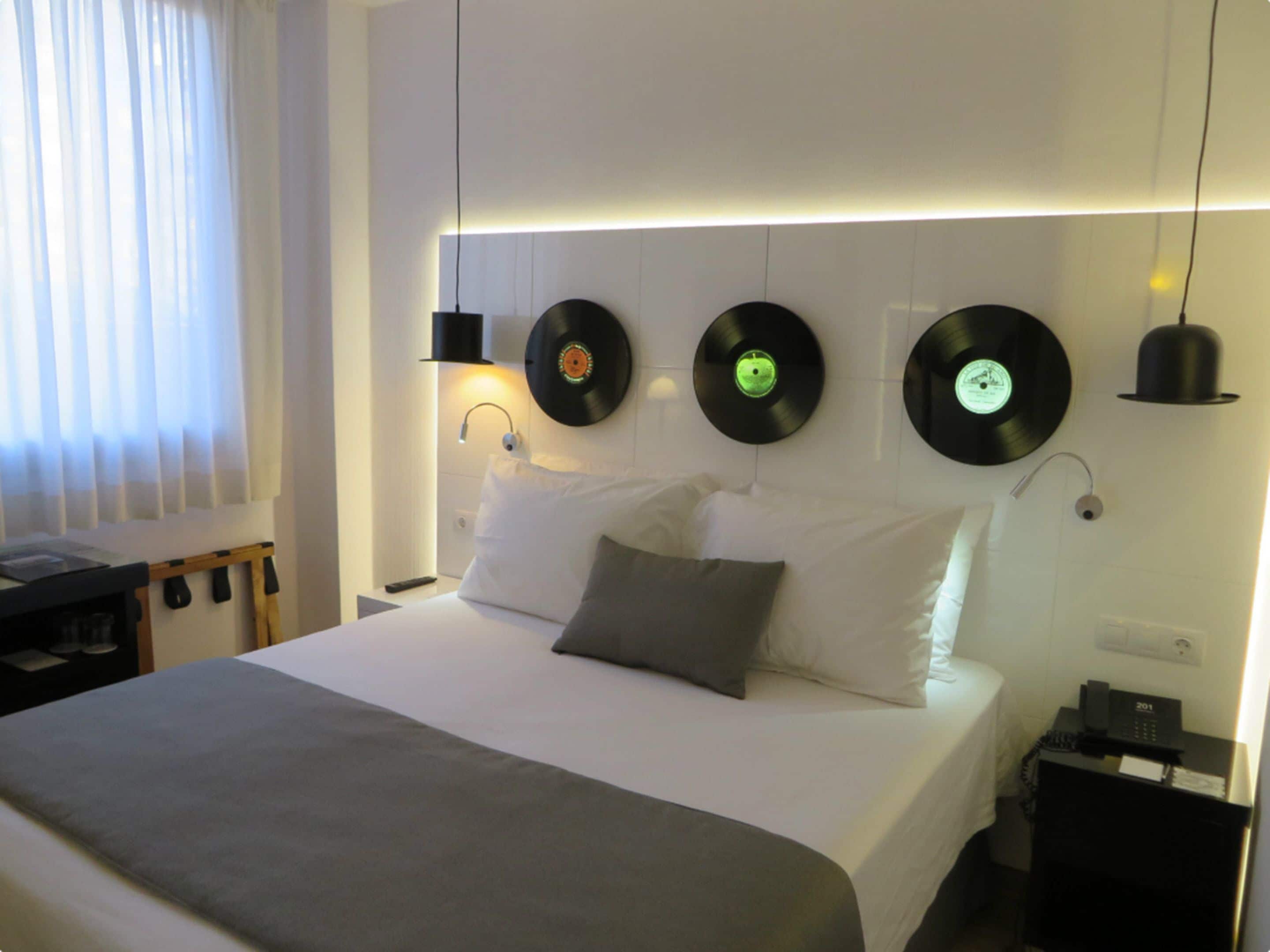 Una habitación de hotel moderna con una cama bien tendida, una mesita de noche con lámpara y tocadiscos montados en la pared.
