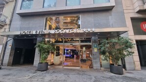 The entrance of Hotel Evania Rocafort with a modern facade, large glass windows, and two potted plants at the entrance. - Evenia Rocafort (Barcelona)