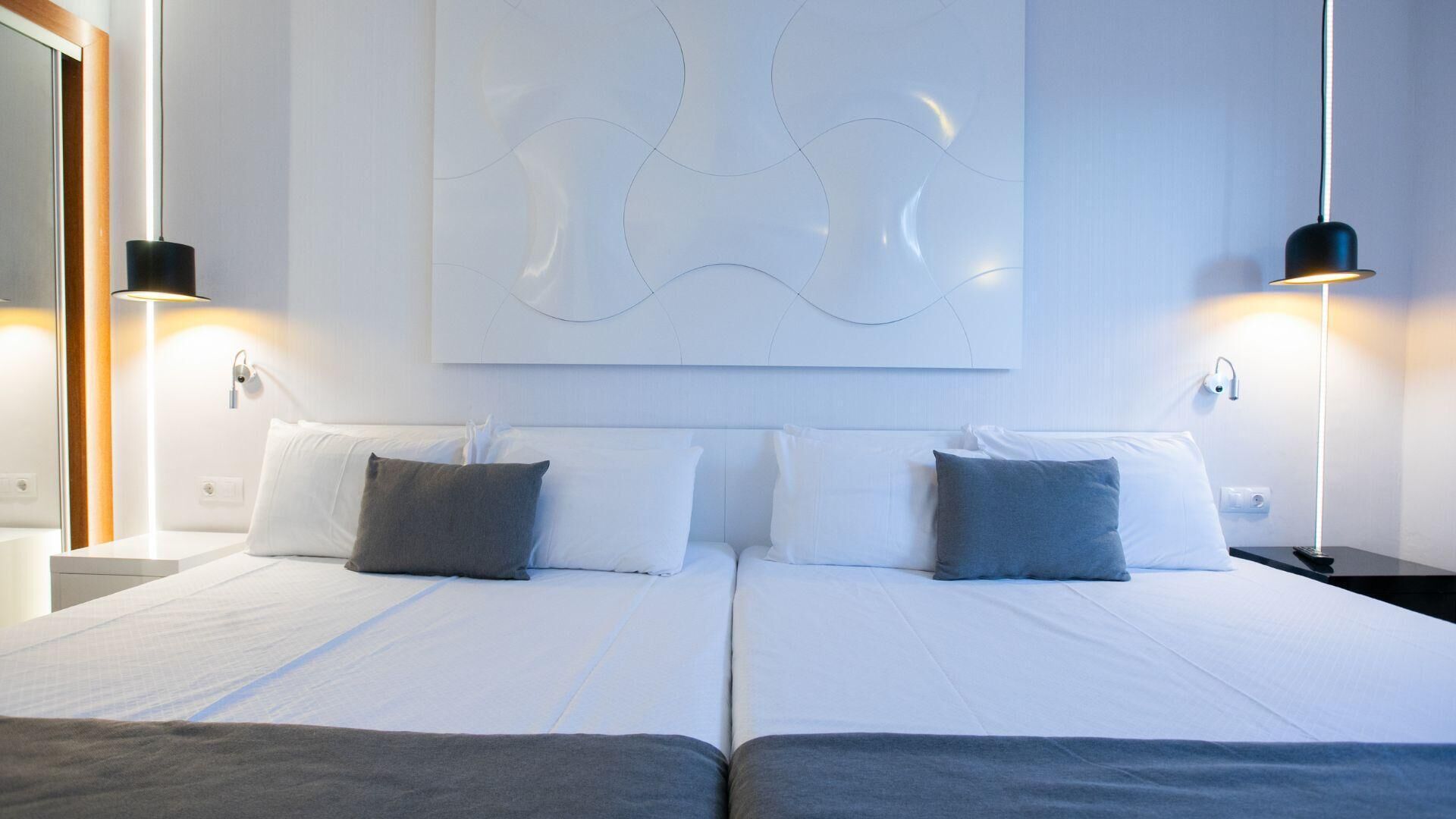 A double bed with white linens and two gray pillows, a wall-mounted abstract art piece, and two black wall sconces.