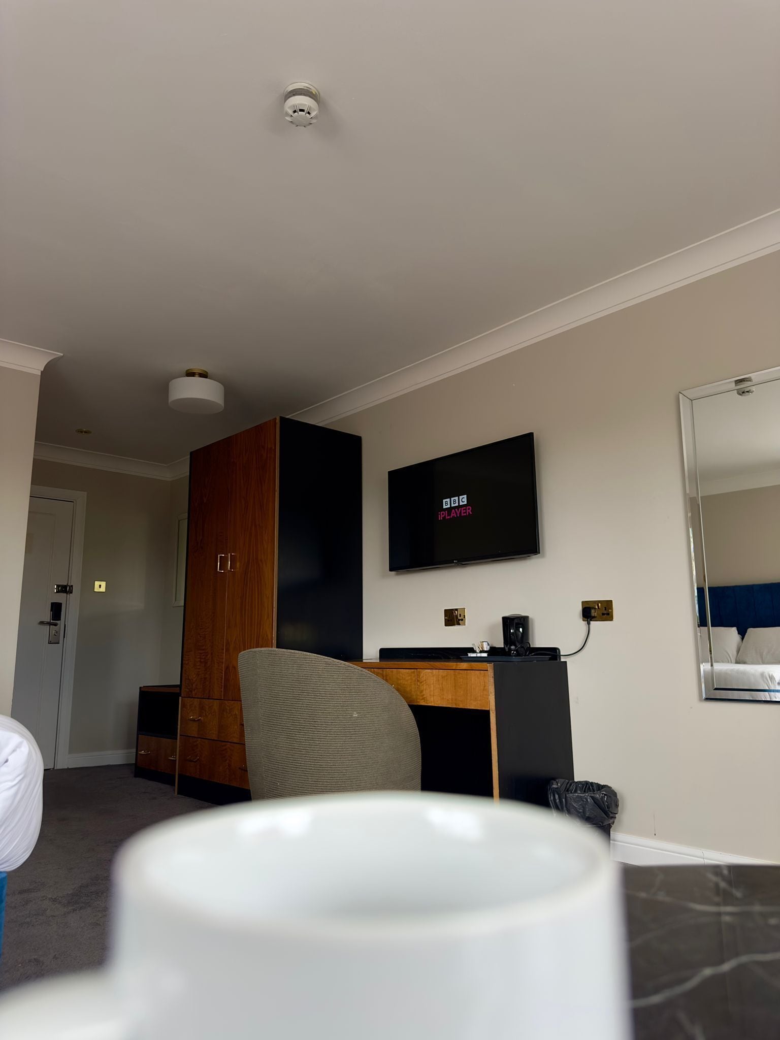 A hotel room with a desk, a television mounted on the wall, a wardrobe, and a mirror reflecting the bed.